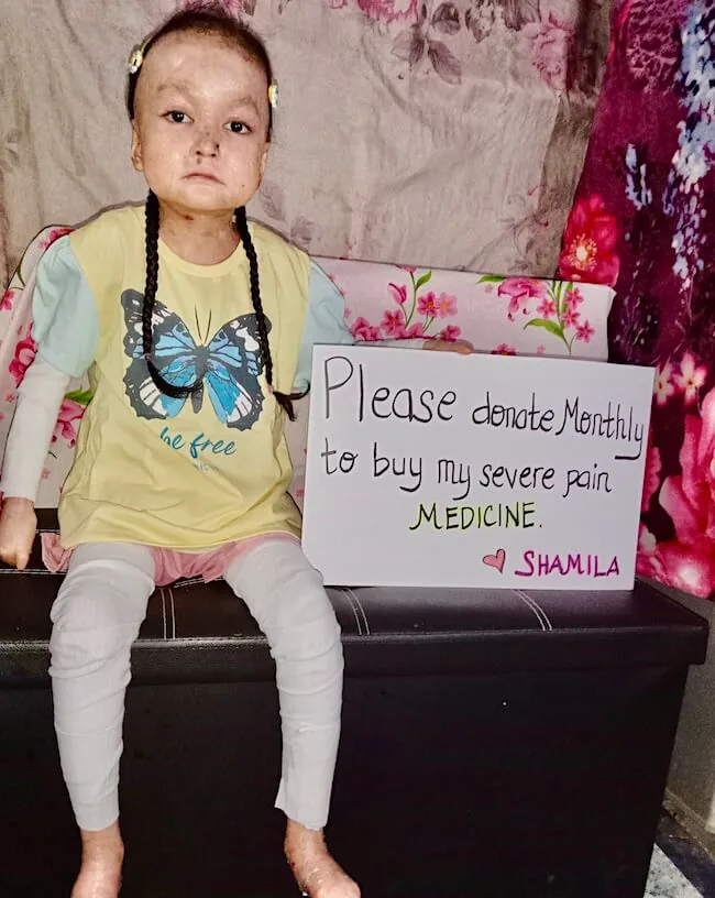 Shamila sitting on a bench holding a handwritten sign that reads: Please donate monthly to buy my severe pain medicine. Shamila